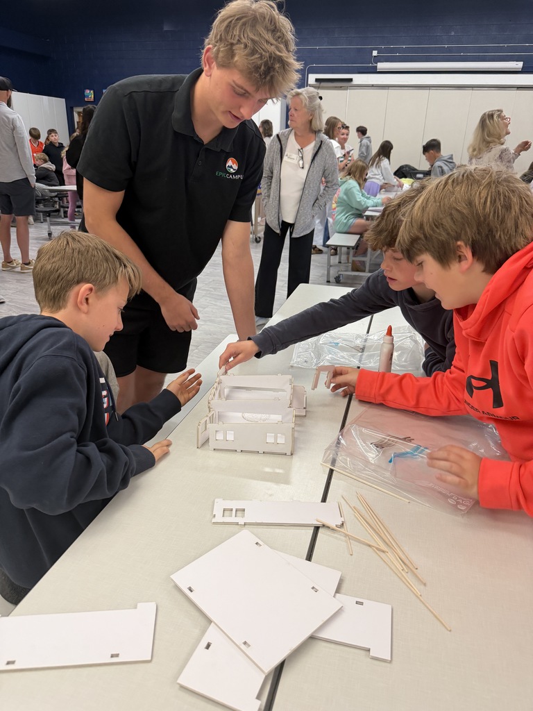A man in a black shirt is standing and helping children in blue and red shirts. They are working on a project at a table with papers and sticks.