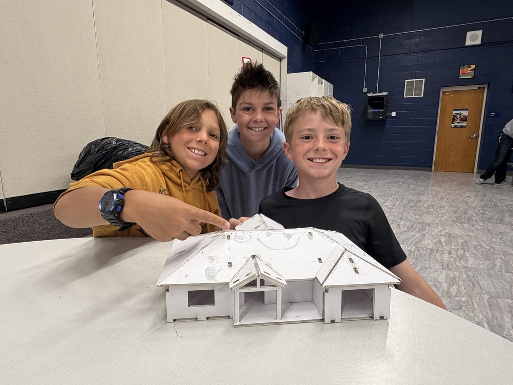 Three children are seated at a table in a room. They are smiling and posing for a photo with a white model house.