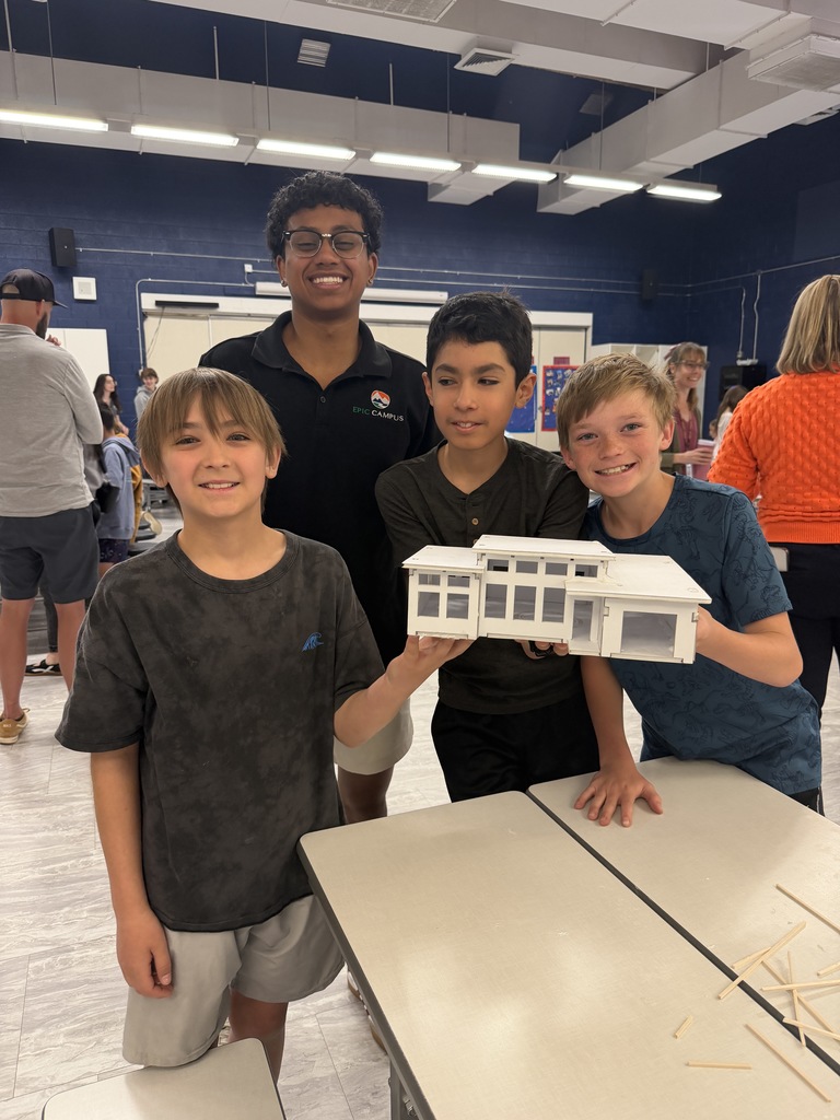 Four boys in a classroom pose with a small architectural model on a table. Bright blue walls and overhead lights are in the background.
