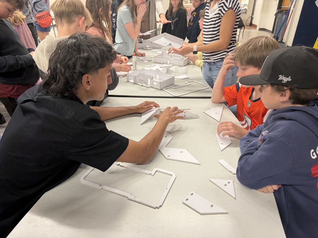 Group of students gathered around a table, working on a project with paper, in a classroom setting.