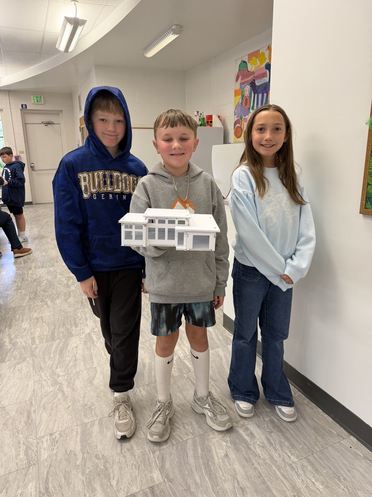 Three children stand in a hallway, two boys and a girl, with one holding a model house.