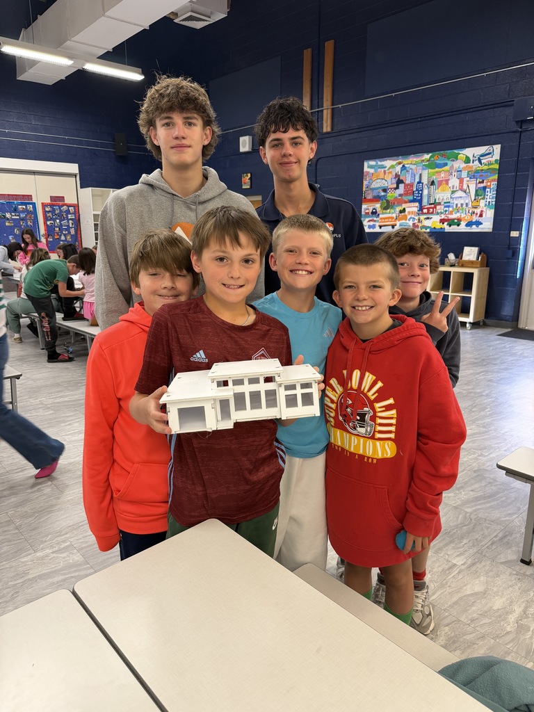 Children pose with a white model structure in a classroom with blue walls, white tables, and a blue floor.