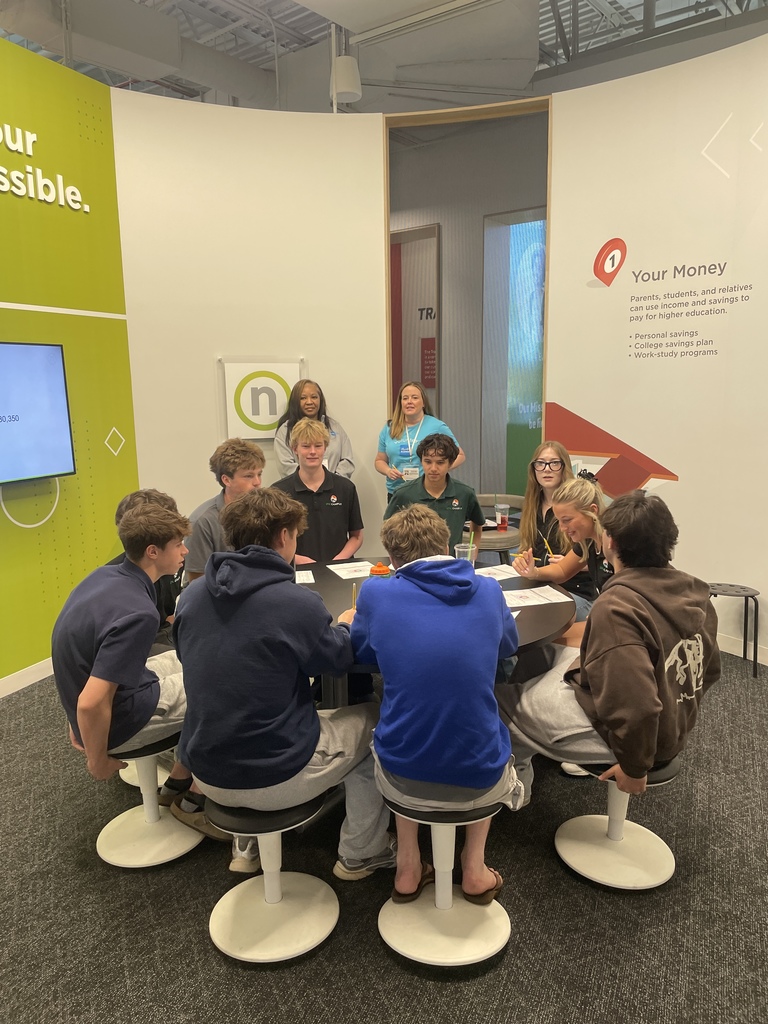 Several individuals sit around a table in a meeting, with one person standing at the back.