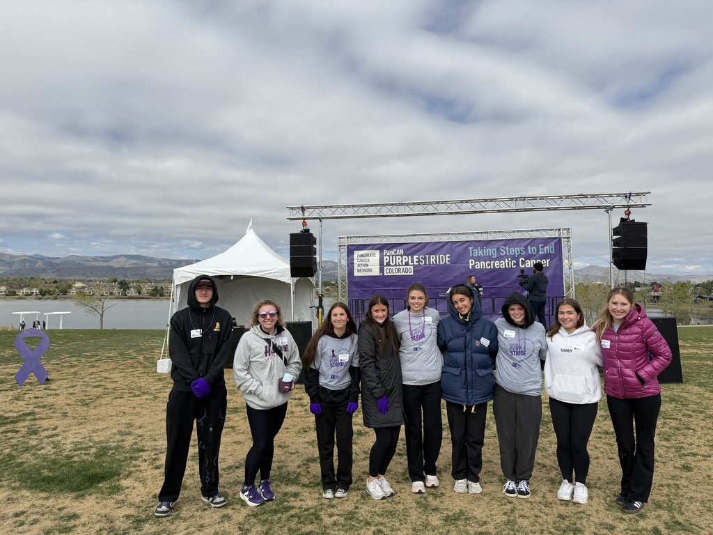 Seven women stand together in a field, with a lake and sky in the background. They wear matching shirts and different colored coats.