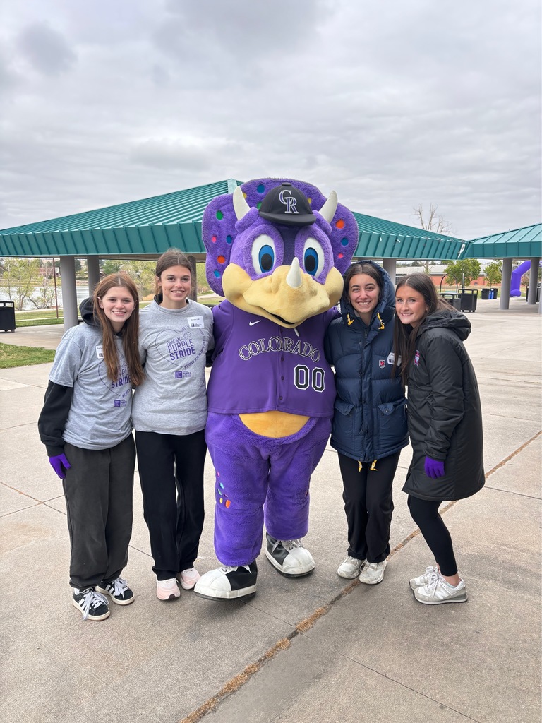 Five people stand near a mascot; four wear jackets, and one wears a purple costume. The group stands outside with a covered area and trees in the background.