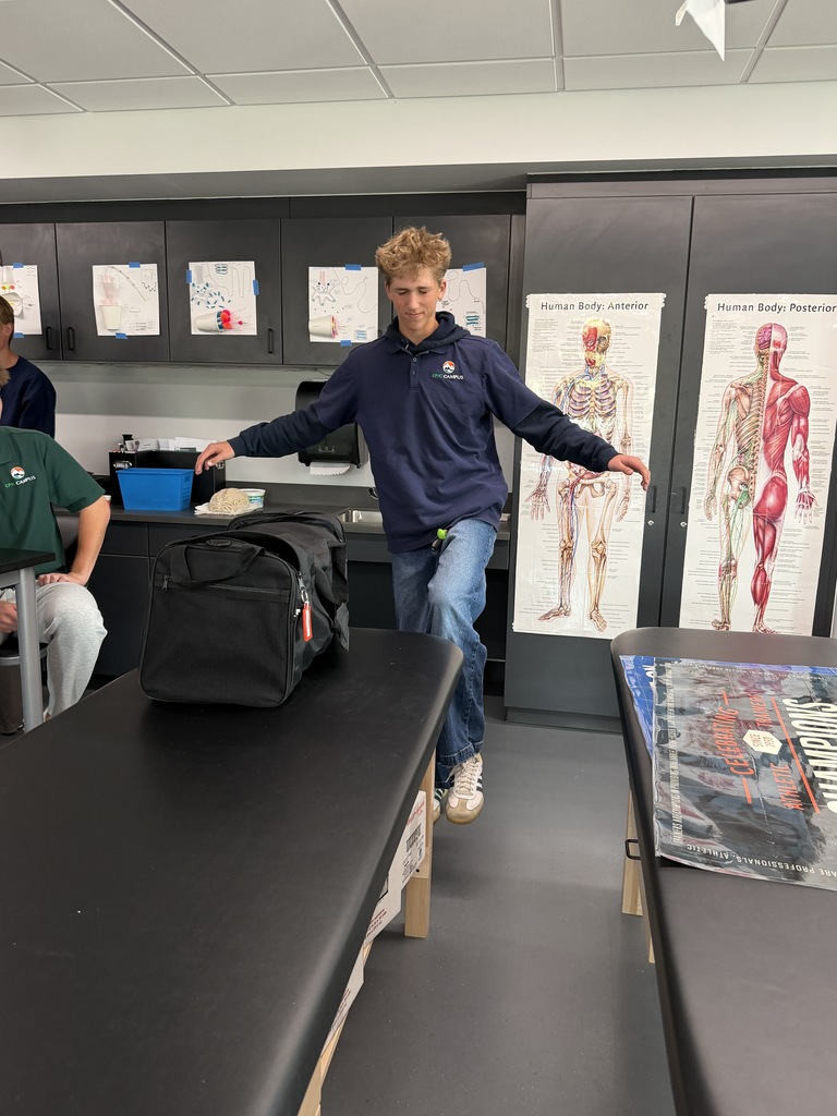 A man stretches on a table in a room with anatomical posters. Another person sits on a chair nearby.