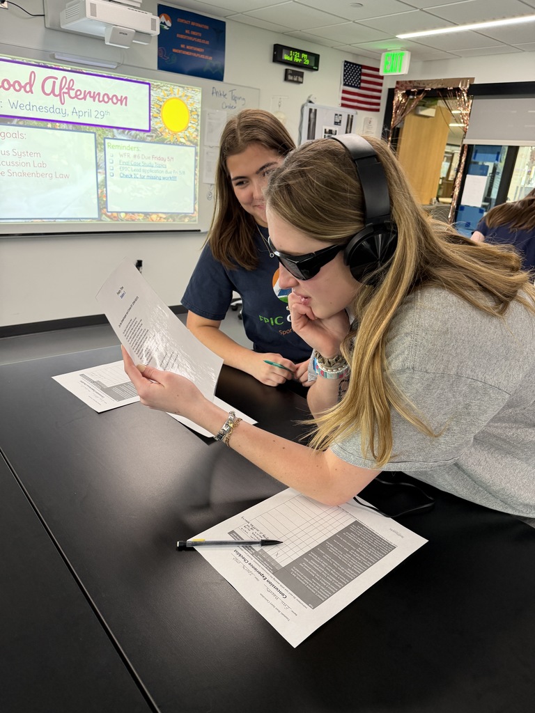 Two women seated at a table, one wearing sunglasses and headphones, reading a document. Behind them, a classroom with a whiteboard.