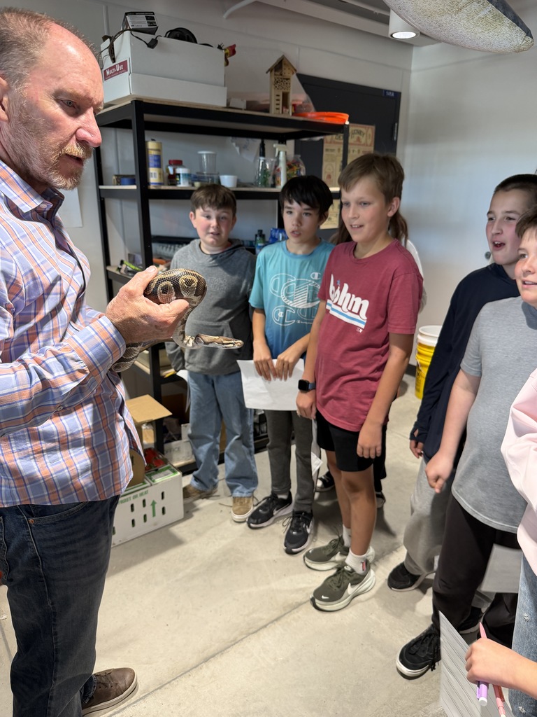 A man holds a reptile as a group of children stand around him in a room with shelves.