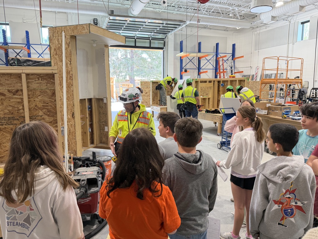 Group of children in a workshop, observing a person in a hard hat and safety vest explaining something.