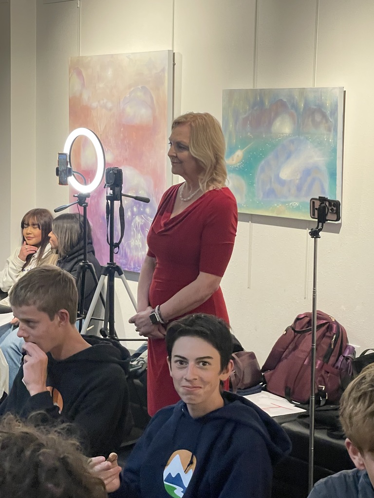 A woman in red stands before a group of people in a room with abstract art. She has a tripod and microphone. Some people are seated with backpacks on the floor.