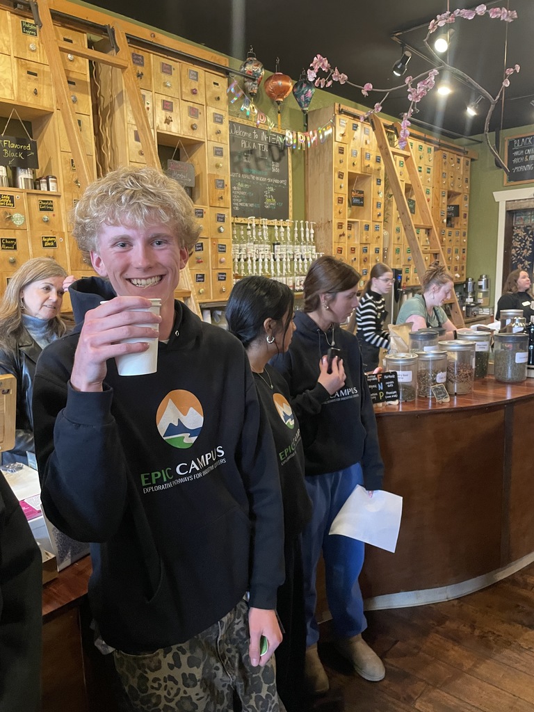 A man holds a cup, smiling. Behind him, people stand in a coffee shop, near a counter and shelves.