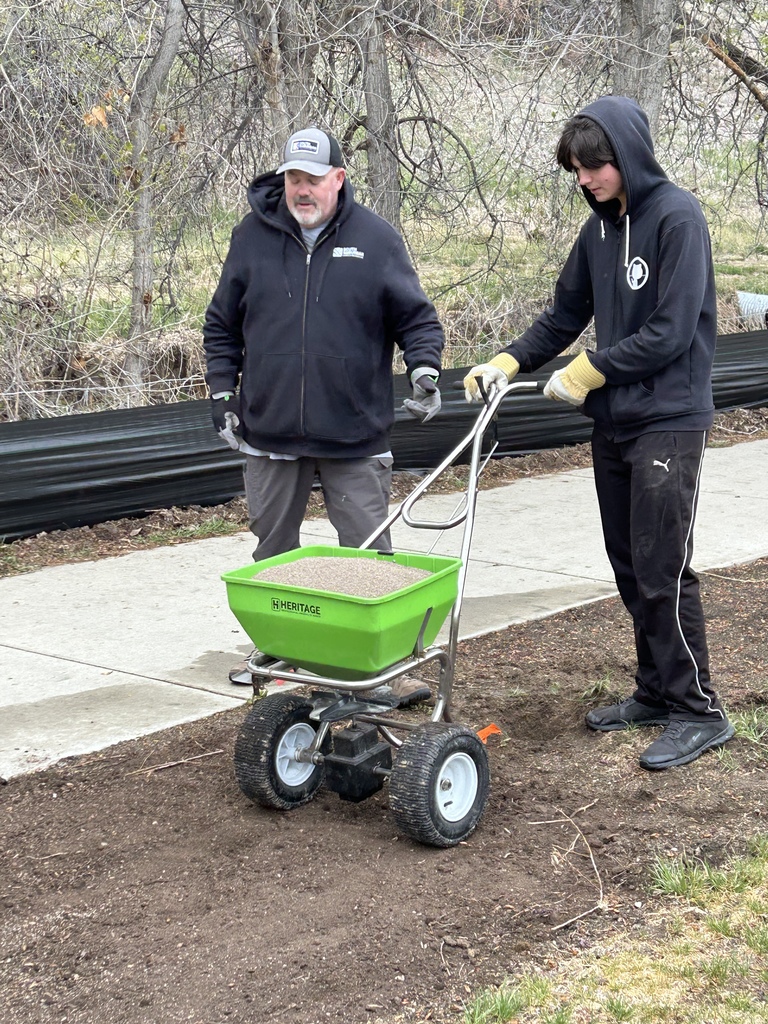 Two individuals in winter gear use a green spreader to add material to a dirt area.