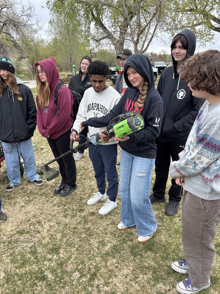 Group of students in various jackets and pants outdoors, one holding a power tool, dry grass as background.