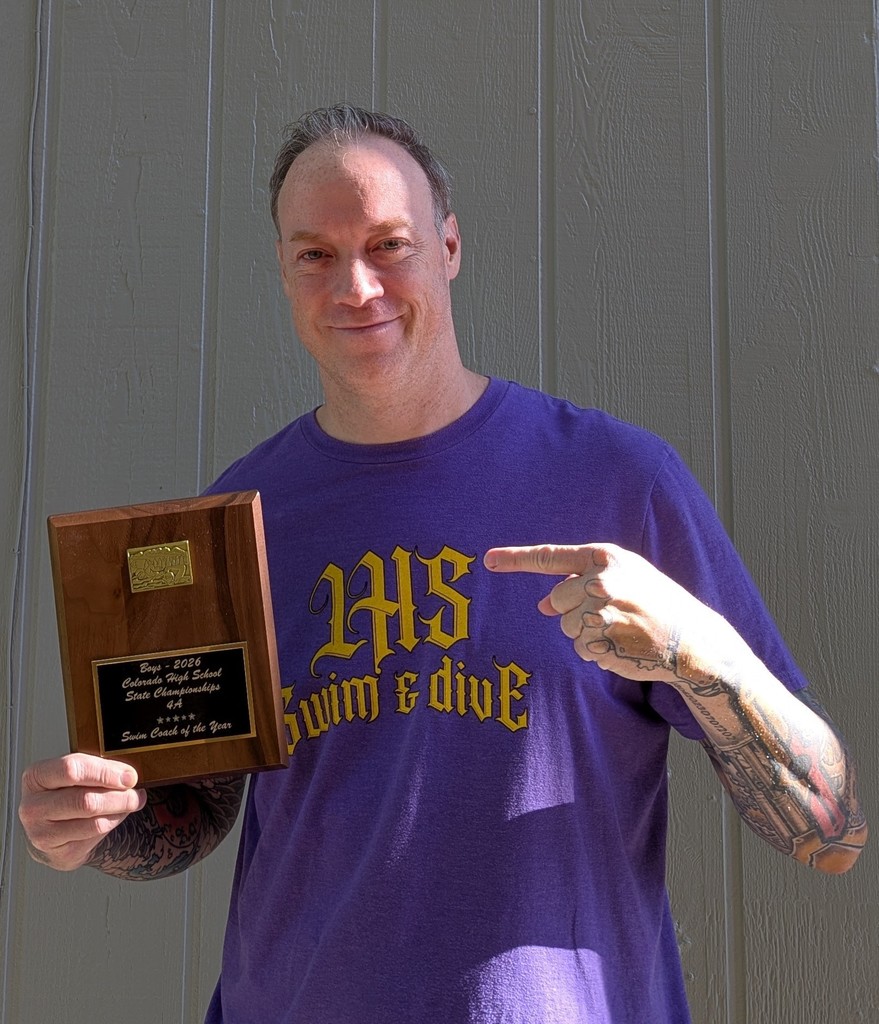 Head boys swim & dive coach Andy Farner wearing purple t-shirt holds plaque for coach of the year, pointing at it. Gray background.