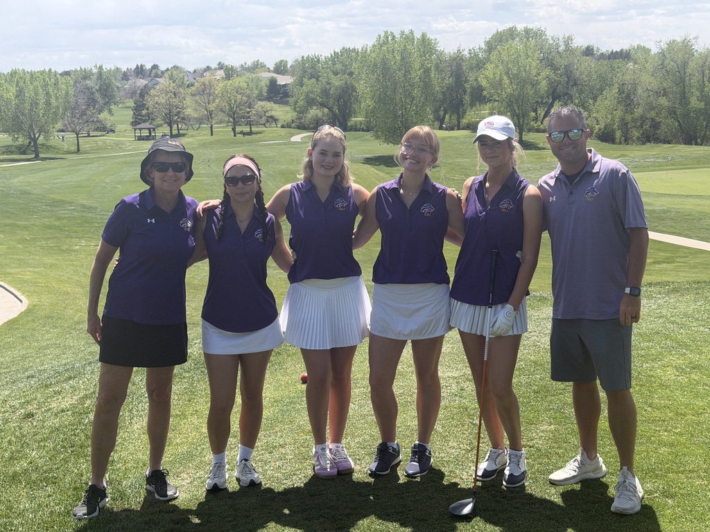 LHS girls golf team and Coach Prince and Coach Walker, in matching purple shirts and white skirts, stands on a golf course. Behind them, a landscape of green trees and hills.