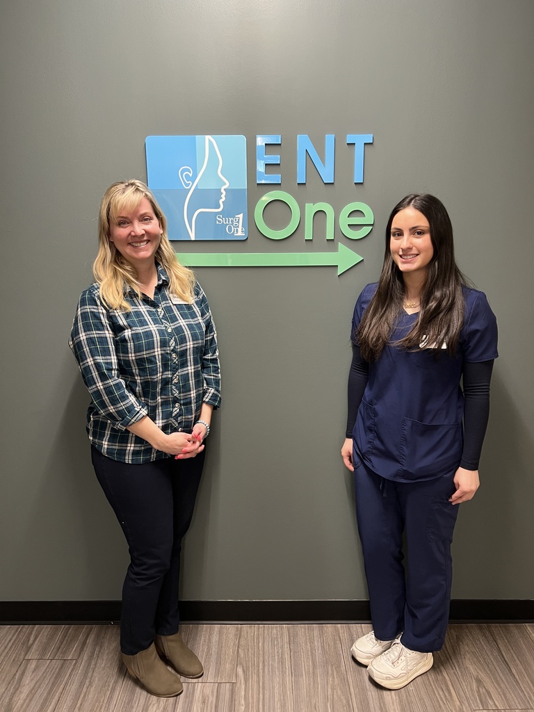 Two women stand next to a sign reading "ENT One" in a clinic setting. One wears a plaid shirt, and the other wears a blue scrub.