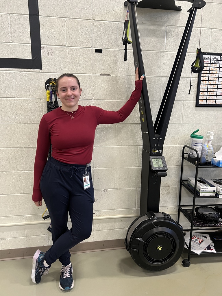 A woman in red top and dark pants stands next to a large black exercise machine.