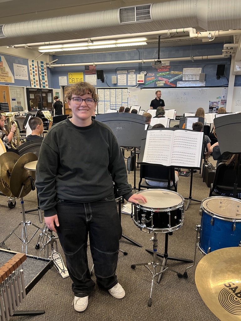 Person in glasses stands next to a drum set in a music room. Many instruments and music stands are visible.