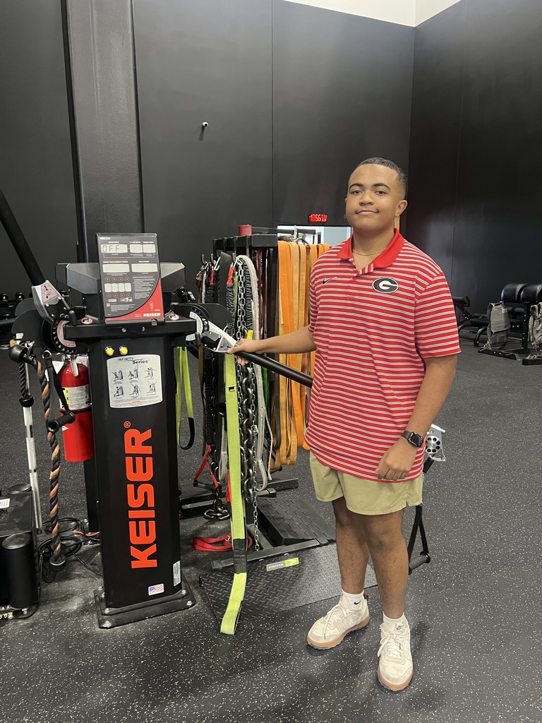 A man wearing a red and white polo, shorts, and sneakers stands near a Keiser weight rack.