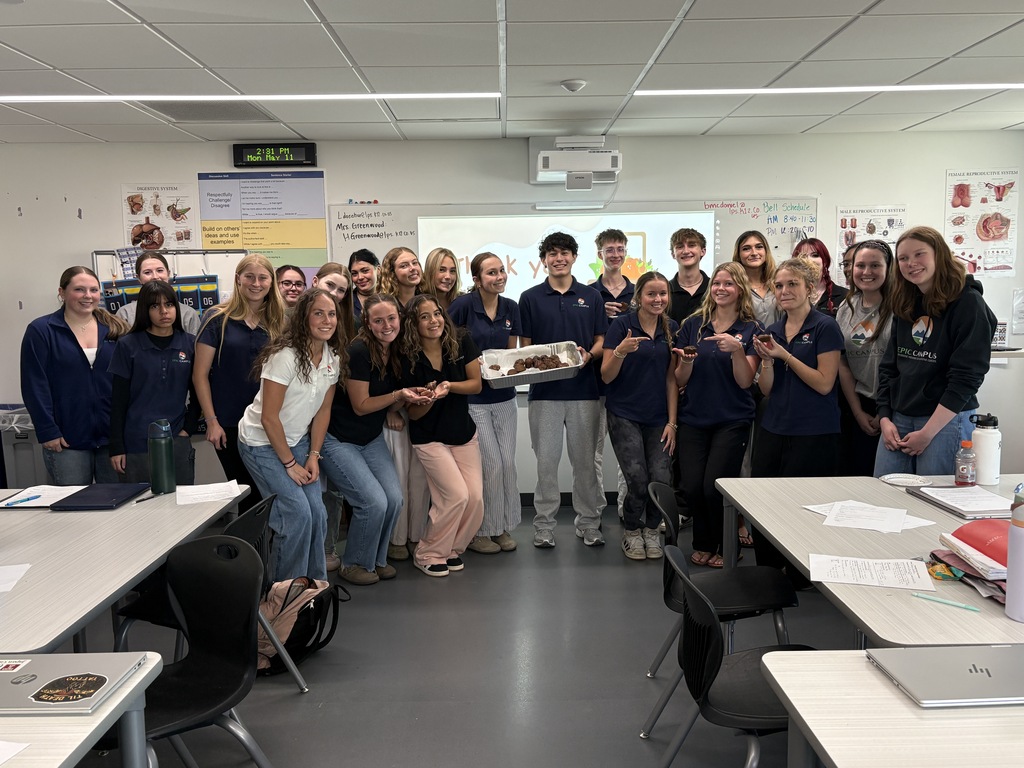 Group of students in blue uniforms posing for a picture in a classroom. Desks and chairs are around them.