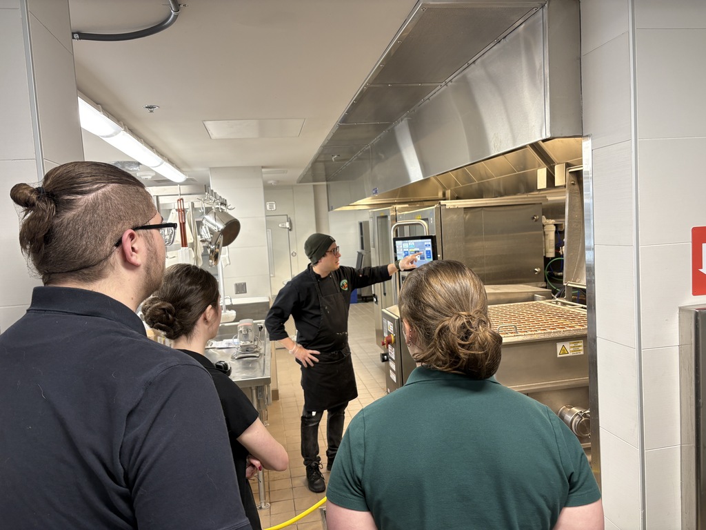 Four people stand in a kitchen. One person wears a green shirt and points to a tablet.