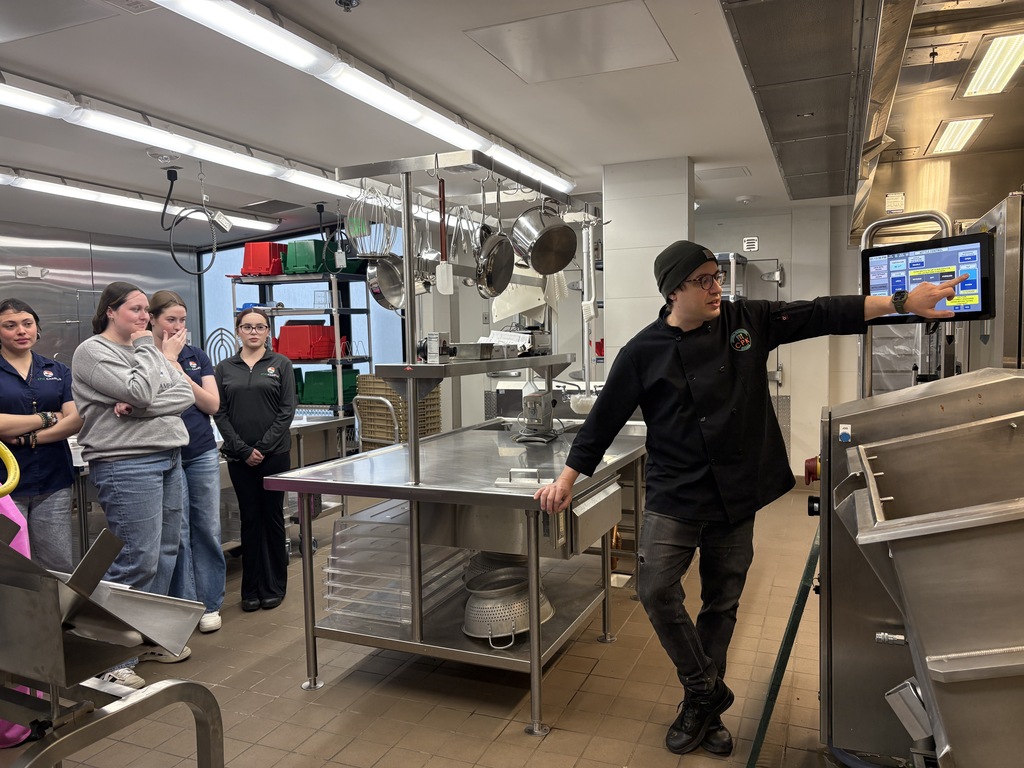 A man wearing a black hat and sneakers points to a monitor in a commercial kitchen. Behind him, people watch, including one in a pink shirt.