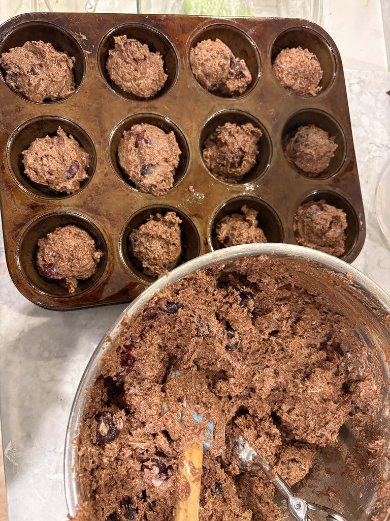 A metal muffin pan with 12 spots filled with brown batter. A bowl nearby contains additional brown batter.