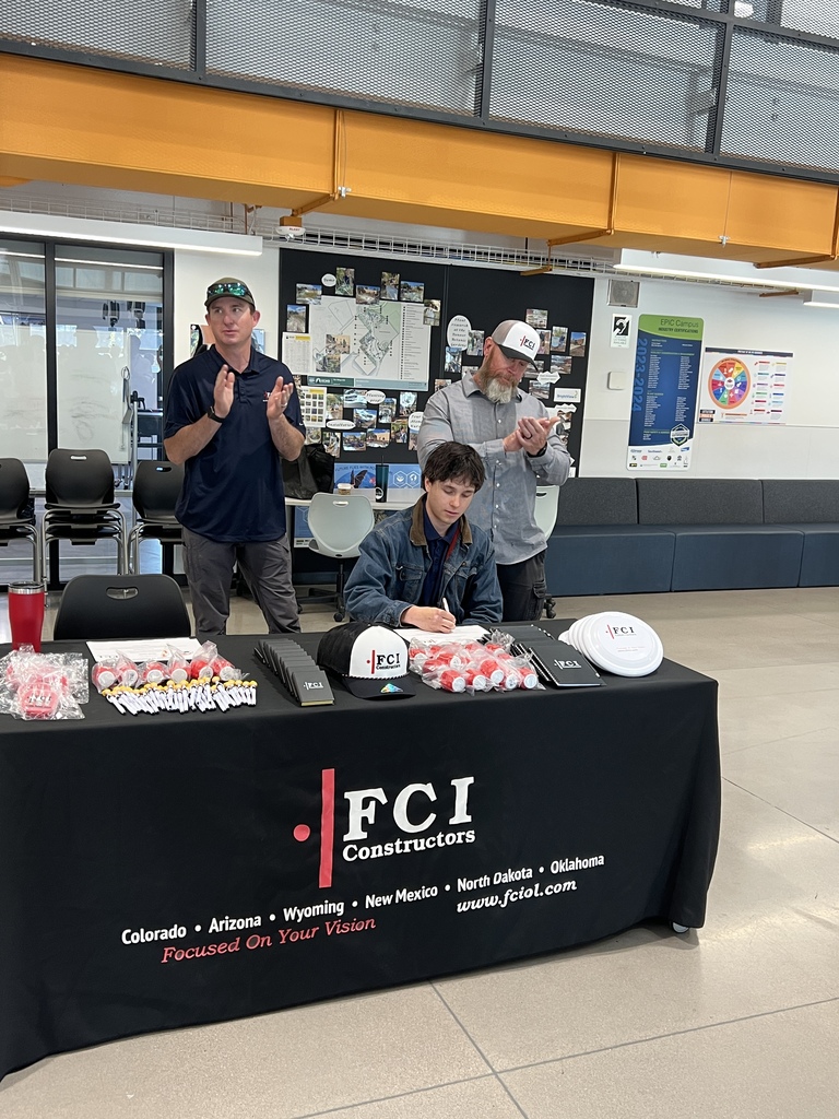 Three people stand behind a table with "FCI" printed on it, with items on the table and a couch behind them.