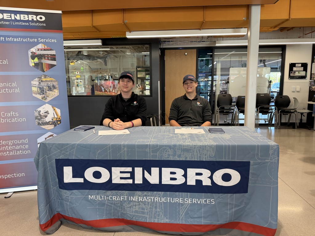 Two people sit at a table covered with a "Loenbro" banner. They wear black shirts and hats.