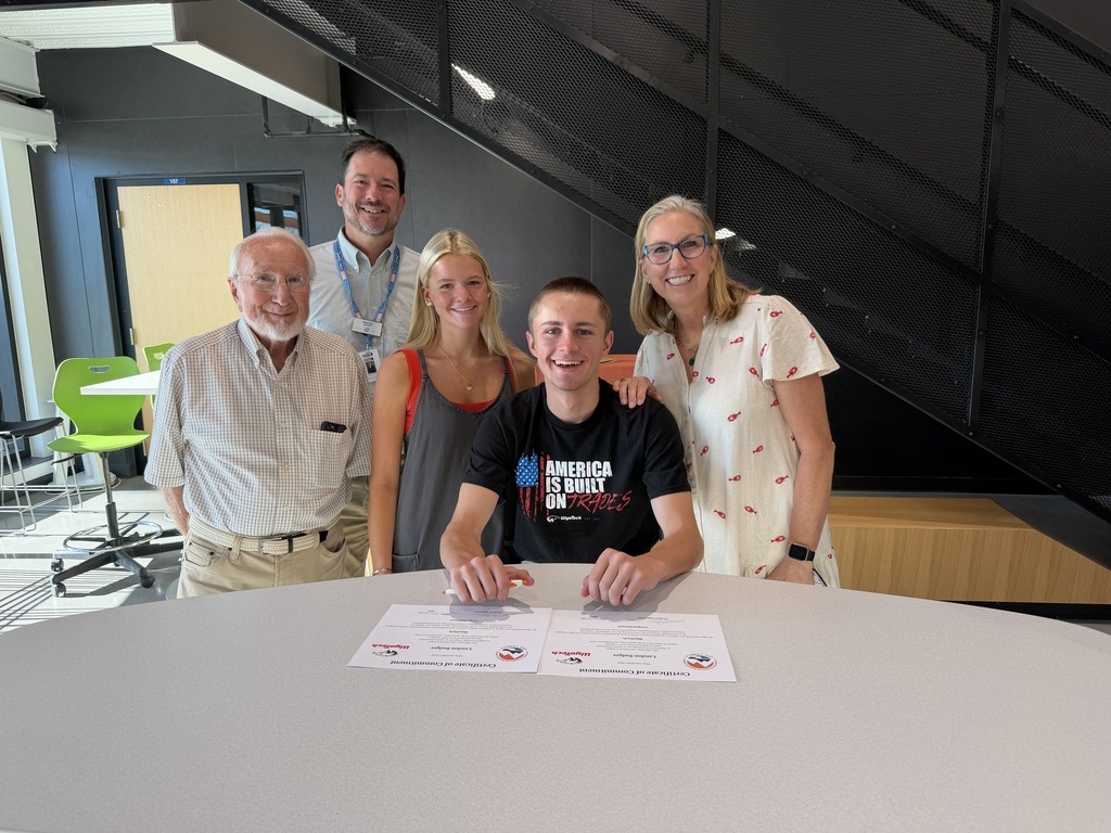 Five people stand around a table in a room, with two holding papers and one signing them.