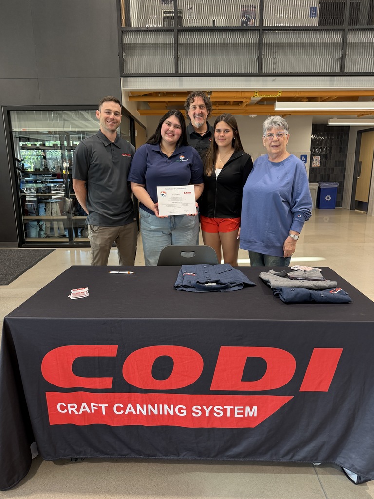Five people stand by a table with a tablecloth that reads "CODI CRAFT CANNING SYSTEM" in red.