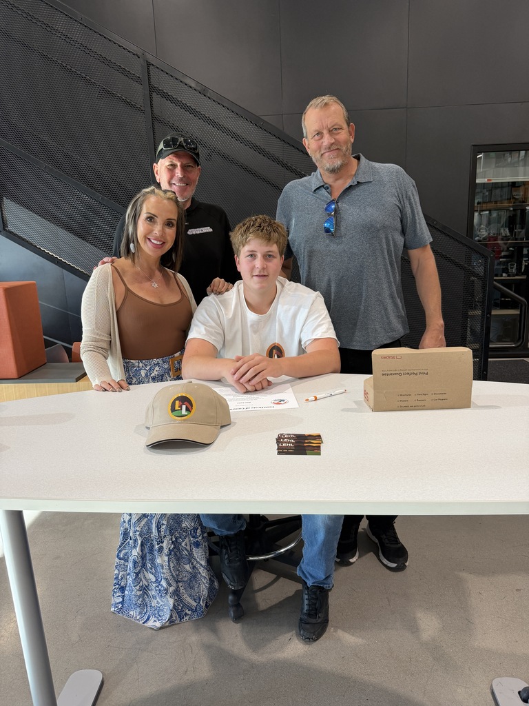 Four people stand behind a table with a box, cap, and other items. A woman stands on the left, a boy on the right, and two men in the middle.