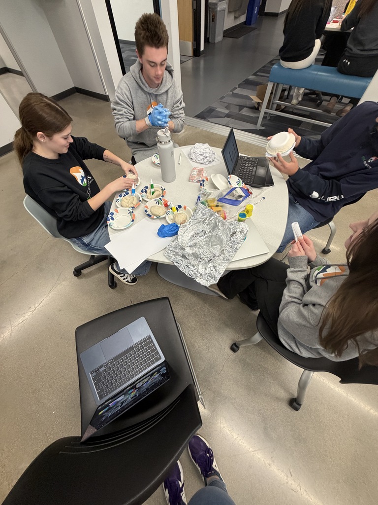 Four people sit around a round table, three wearing hoodies. They work on tasks at the table, such as icing a cupcake.