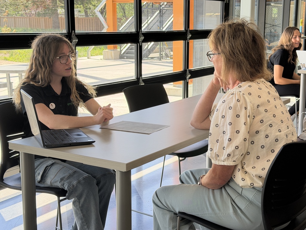 Two individuals sit at a table with a laptop, one wearing glasses. Behind them, a staircase leads to a glass wall.