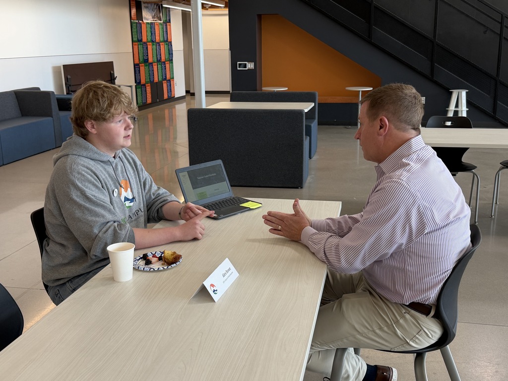 Two men engage in conversation at a table with a laptop, one wearing glasses and the other in a belt.