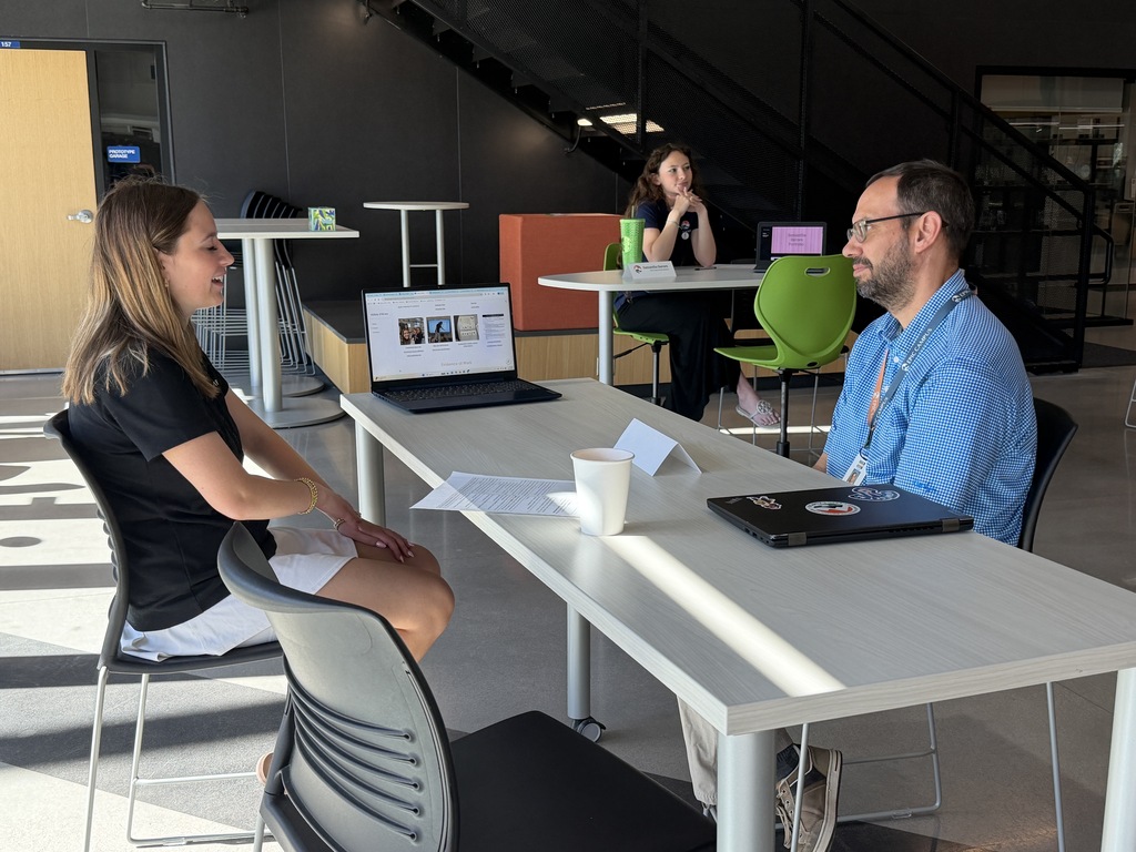 Two people in an office setting have a conversation. One person sits on a chair, the other on a stool.