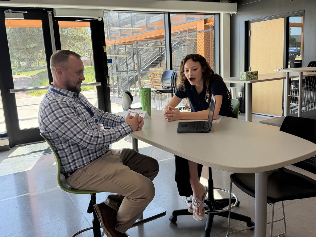 A man and a woman sit at a table, engaging in conversation while a laptop rests between them.
