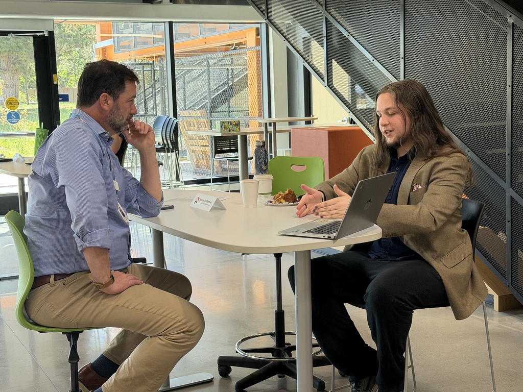 Two people sit at a table with a laptop, discussing. One wears a blue shirt, the other a brown jacket.
