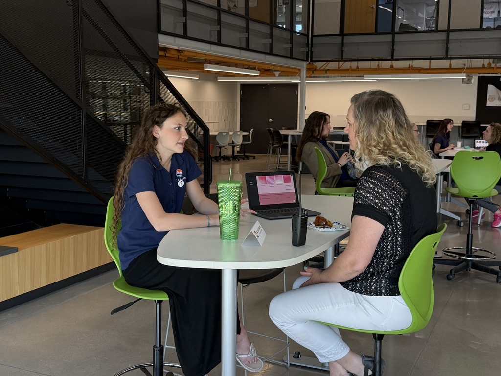 Two women in a modern office with a table between them. One wears a blue shirt, the other a black top.