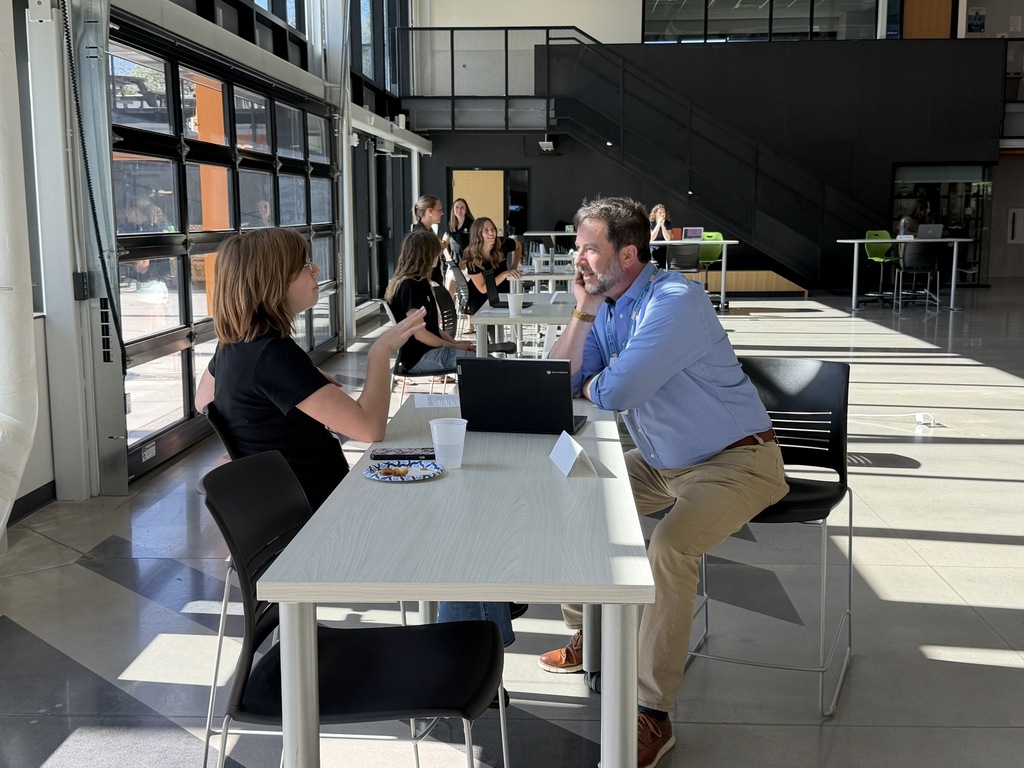 Two people sit at a table in a modern room. A man speaks while the woman listens.
