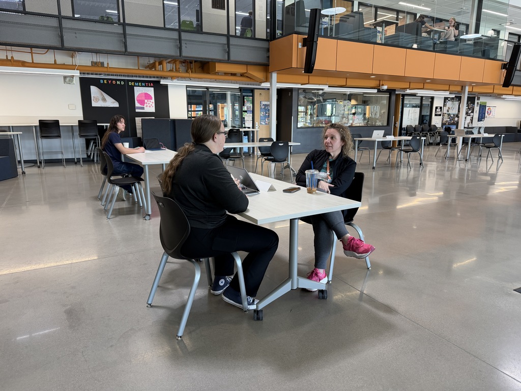Two people seated at a desk in a spacious, open-plan office area with chairs, tables, and large windows.