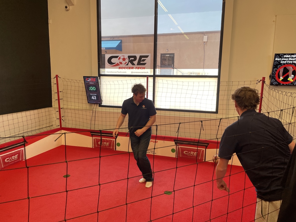 Two men in a gym with a red floor and a large net, playing a game. A wall displays "CORE" and a digital sign.