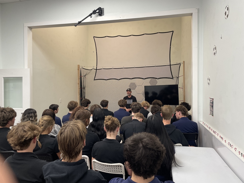 A room with a white backdrop features a large screen and a person standing in front. An audience sits in chairs.