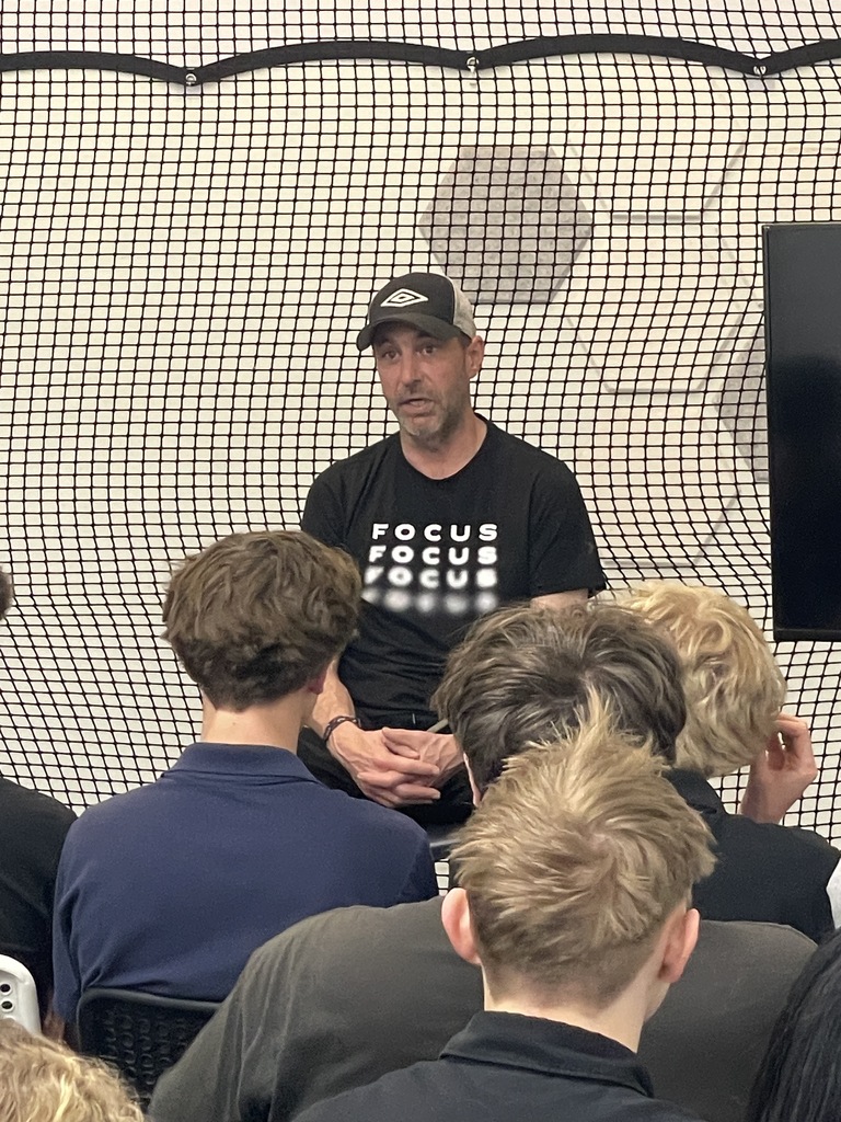 A man in a cap speaks to a group of young people in a room with a black and white net in the background.