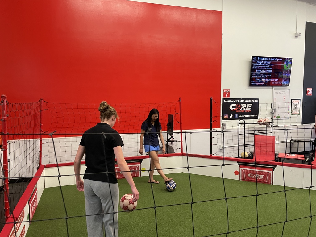 Two women play soccer in a gym with a red wall, white floor, and net, one holding a ball.