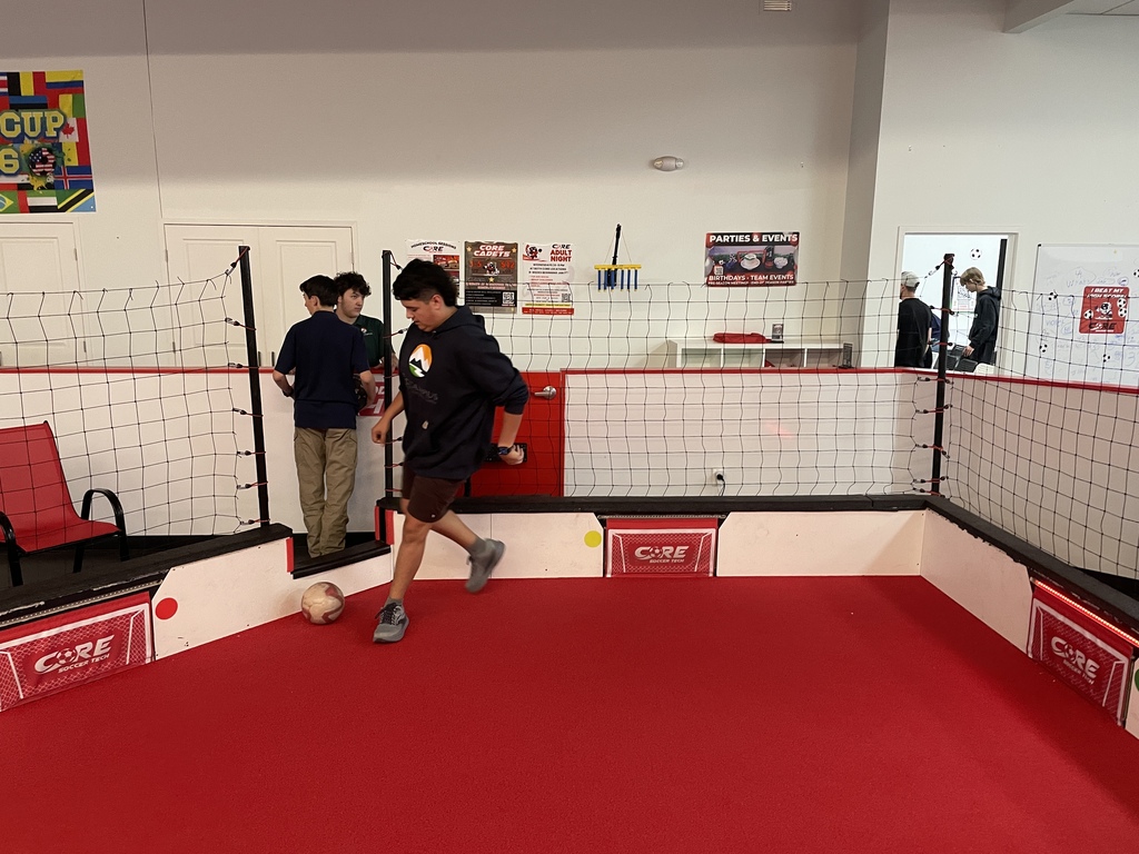 Two people are playing soccer in a red-floored room with a net, with another person standing nearby.