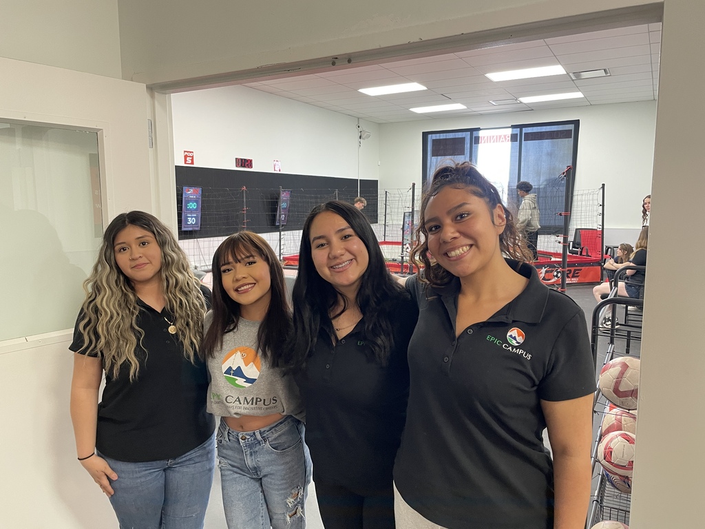 Four women in matching shirts and jeans smile at the camera in a gym with gym equipment and a large window.