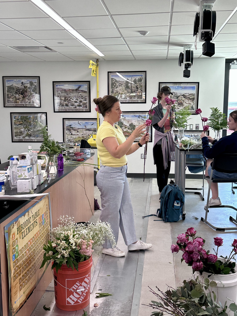 A woman in a yellow shirt holds a flower arrangement. Behind her, a person arranges flowers at a table.
