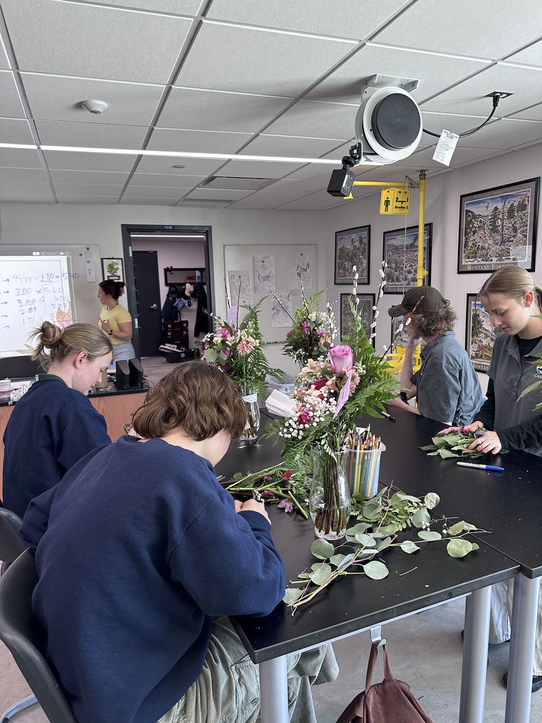 Five individuals working on a floral arrangement at a table in a room with picture frames on the wall.