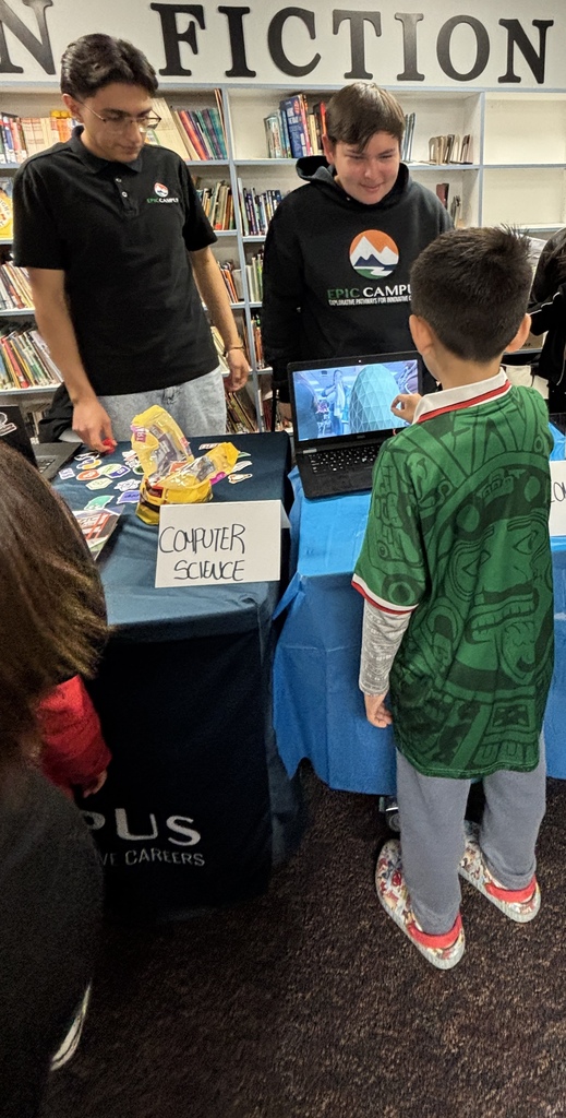 A table with a blue cloth displays a laptop and a sign reading "Computer Science." A boy is looking at the screen, while two men stand behind him. Behind them, a bookshelf has a large sign reading "Non Fiction."